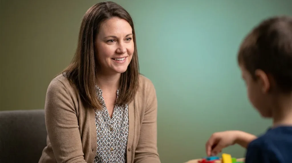 Smiling woman in a tan cardigan seated at a table across from a young boy playing with toys, illustrating what is ABA therapy